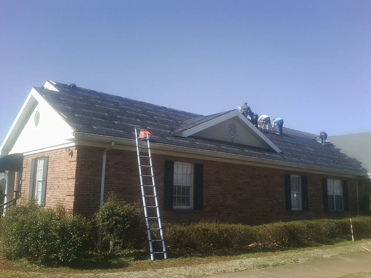 Crew tearing shingles off a brick ranch roof with a ladder set — Emergency Roof Repair work by our crew in Disputanta